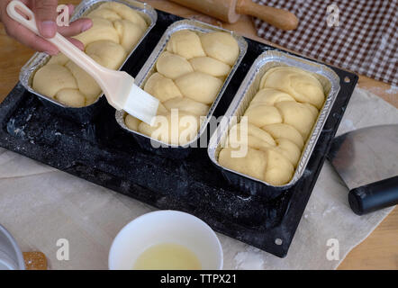 Cropped hand of baker applying egg white with brush on bread in container Stock Photo