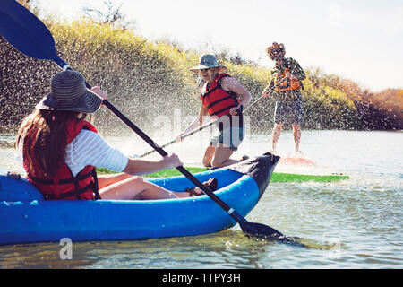 Friends splashing water on woman sitting in inflatable kayak while paddleboarding at lake Stock Photo