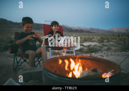 Happy brothers eating marshmallows while sitting on chairs at campsite during dusk Stock Photo