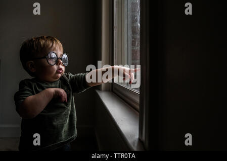 Toddler boy wearing round glasses standing by window pointing outside Stock Photo