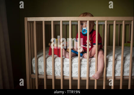 Portrait of baby boy sucking pacifier while sitting in crib at home Stock Photo