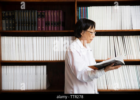Senior female scientist reading book by shelves at laboratory Stock Photo