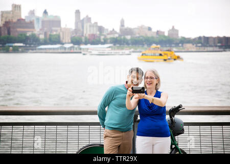 Happy mature couple taking selfie while standing on promenade by river in city Stock Photo