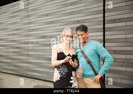Mature couple using smart phone while standing against wall Stock Photo