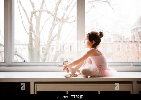 Girl sitting on table and wearing ballet shoes against window Stock Photo