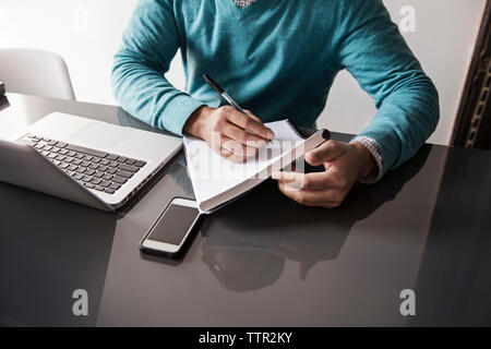 Midsection of man writing on book by laptop at table in office Stock Photo