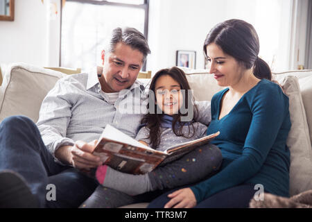 Girl with father and mother reading book while sitting on sofa at home Stock Photo