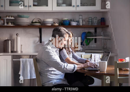 Daughter sitting with father using laptop computer in kitchen at home Stock Photo