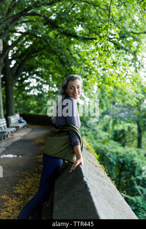 Smiling woman looking away while leaning on retaining wall against trees Stock Photo