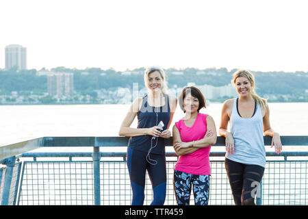 Portrait of confident female friends in sports clothing while standing on bridge against clear sky Stock Photo