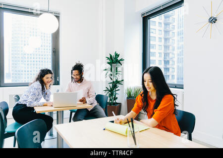 businesswoman reading documents while colleagues discussing over laptop computer in office Stock Photo