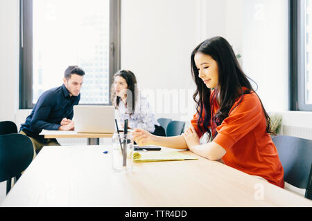 businesswoman examining documents while coworkers discussing over laptop computer in background at office Stock Photo