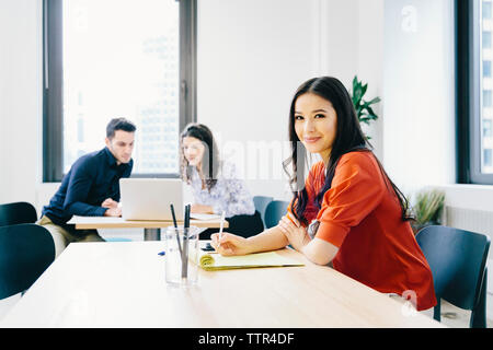 Portrait of businesswoman with documents sitting against colleagues discussing over laptop computer in background at office Stock Photo