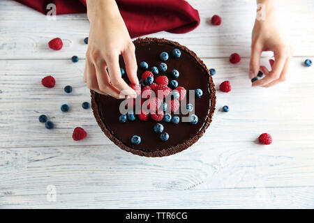 Female hands decorating chocolate tart with berries on wooden background Stock Photo