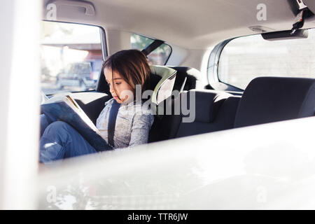 Serious boy reading book while sitting in car seen through window