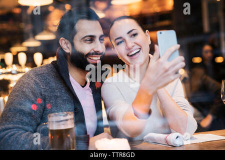 Happy woman with boyfriend using mobile phone seen through restaurant window Stock Photo