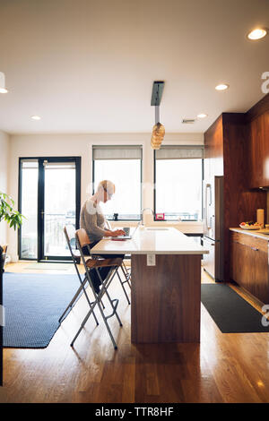 Side view of senior woman using laptop while sitting at kitchen table Stock Photo
