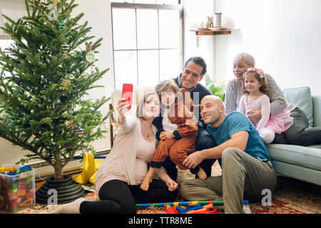 Pregnant woman taking selfie while sitting with family by christmas tree at home Stock Photo
