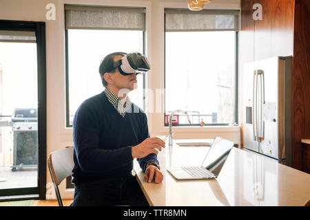 Man wearing virtual reality simulator sitting at table in kitchen Stock Photo
