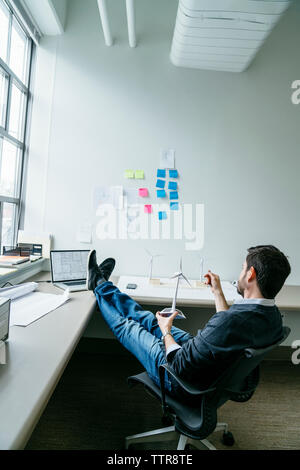 Side view of businessman holding wind turbine model while sitting in office Stock Photo