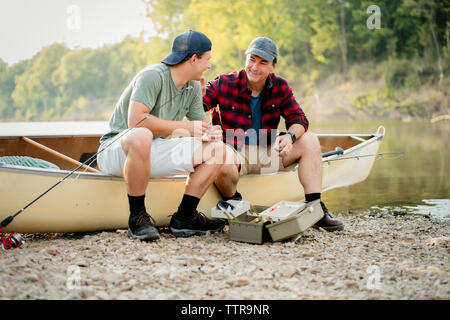 Happy friends talking while adjusting fishing tackles on boat at lakeshore Stock Photo