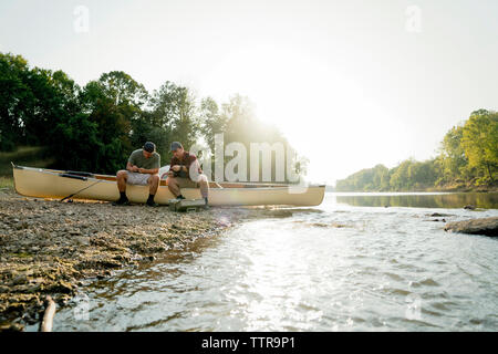 Male friends adjusting fishing tackles while sitting on boat at lakeshore against clear sky Stock Photo