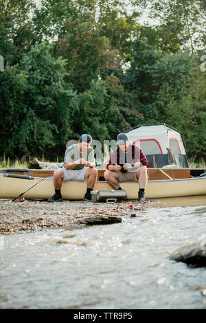 Friends adjusting fishing tackles while sitting on boat by lake at campsite Stock Photo