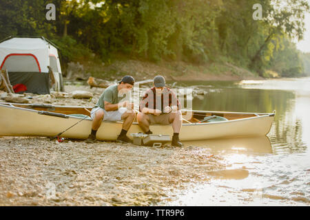 Friends adjusting fishing tackles while sitting on boat at campsite Stock Photo