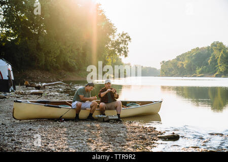 Male friends adjusting fishing tackles while sitting on boat at campsite by lake Stock Photo