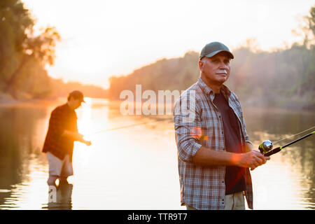 Senior man looking away while friend fishing in background during sunset Stock Photo