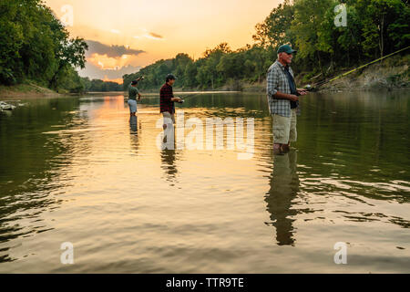 Friends enjoying fishing in lake against sky during sunset Stock Photo