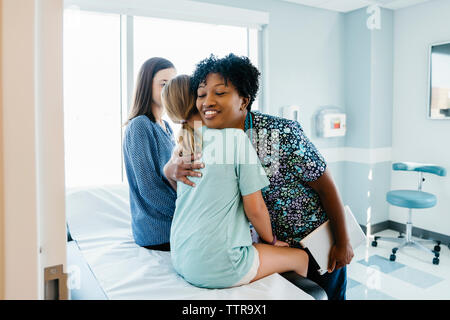 Pediatrician embracing girl sitting by mother on examination table in hospital Stock Photo