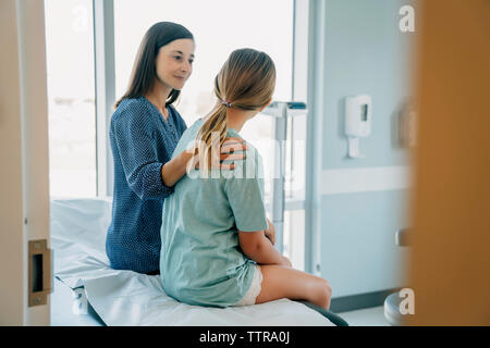 Mother consoling daughter while sitting on examination table in hospital seen through doorway Stock Photo
