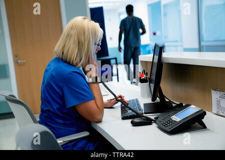 Female doctor talking on telephone while using desktop computer with male colleague in background Stock Photo