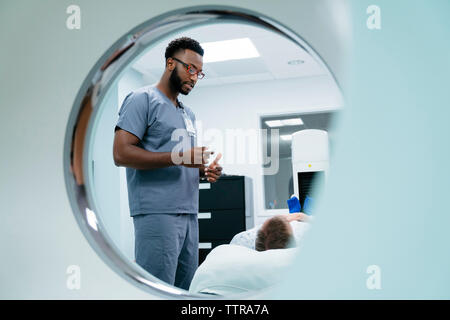 Male nurse talking with patient lying in examination room seen through MRI Scanner Stock Photo