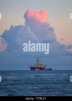 Drilling rig in sea against cloudy sky during sunset