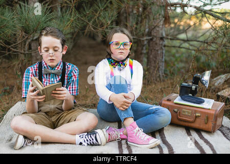 Siblings in old-fashioned costume sitting on blanket with antique camera at forest Stock Photo