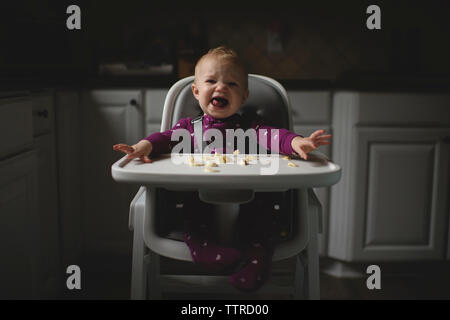 Portrait of cute baby girl crying while sitting on high chair in kitchen at home Stock Photo