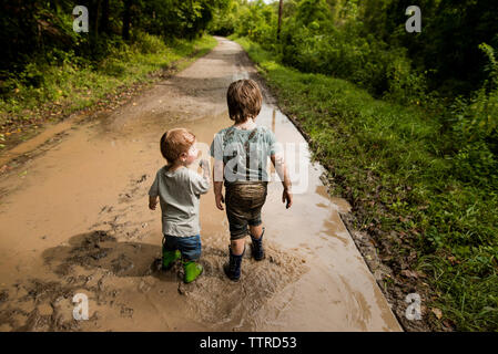 Rear view of messy brothers walking in dirty puddle on road Stock Photo