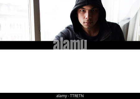 Portrait of young man wearing black hooded jacket sitting by window at home Stock Photo