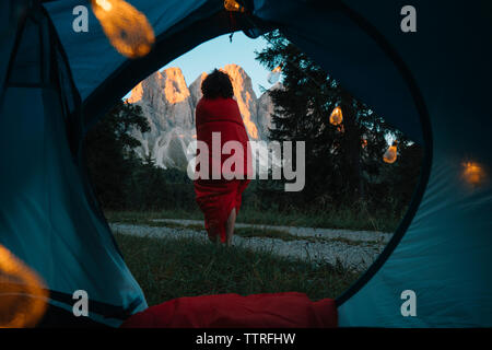 Rear view of young woman standing against mountains while being wrapped in blanket seen through tent Stock Photo