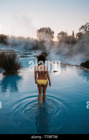 Rear view of woman wearing bikini while walking in thermal pool during sunset Stock Photo