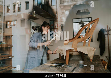 Female upholsterer making wooden chair on table in workshop seen through window Stock Photo
