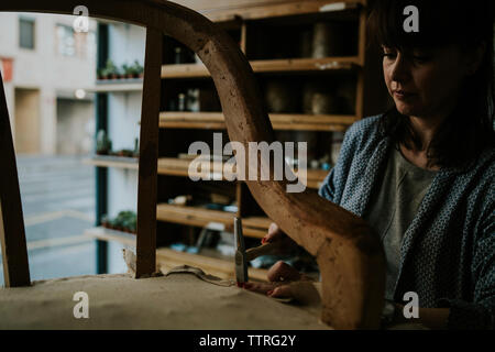 Confident female upholsterer using hammer while upholstering wooden chair in workshop Stock Photo