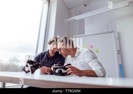 Technicians examining virtual reality simulators while sitting by window at table Stock Photo