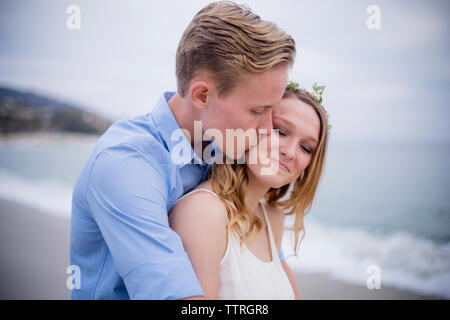 Man kissing girlfriend while standing at beach against sky Stock Photo