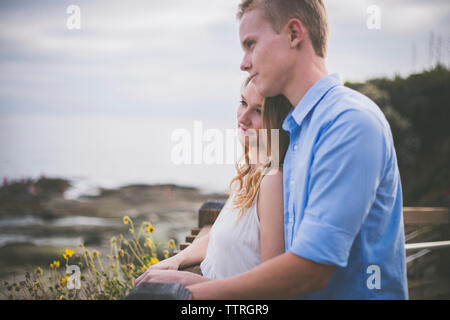 Smiling couple looking away while standing by railing against cloudy sky Stock Photo