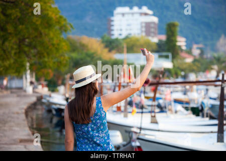 Rear view of woman wearing hat taking selfie while standing at harbor in town Stock Photo