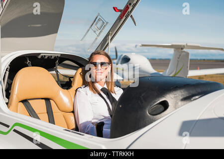 Portrait of confident female pilot wearing sunglasses sitting in airplane against blue sky at airport during sunny day Stock Photo