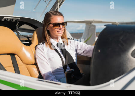 Confident female pilot wearing sunglasses sitting in airplane against blue sky at airport during sunny day Stock Photo